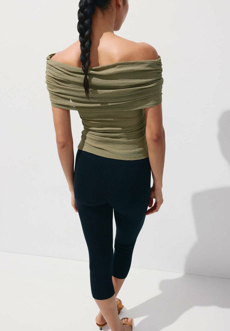Femme aux longs cheveux tressés portant un top vert olive bardot et des leggings capri noirs, debout devant un fond blanc uni.