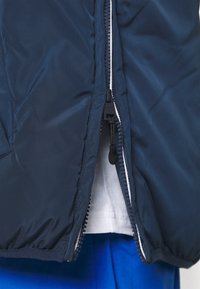 Chaqueta de plumas azul marino con cremallera completa y forro blanco en contraste. Presenta puños elásticos y un exterior suave y brillante.