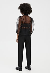 Zwarte doorzichtige blouse met pofmouwen en een stippenstructuur, gelaagd over een zwarte kanten bralette, gecombineerd met zwarte broek en schoenen.