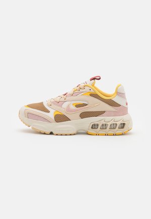 Zapatilla de running Nike en beige, rosa y amarillo. Materiales de malla y ante con una suela intermedia gruesa y bolsas de aire visibles. Punta redondeada y cordones.