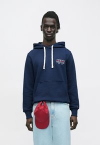 Νέος άνδρας που φοράει μπλε σκούρο φούτερ Tommy Hilfiger και ανοιχτόχρωμα μπλε τζιν με κόκκινο καπέλο περασμένο στη μέση, στέκεται μπροστά σε λευκό φόντο.