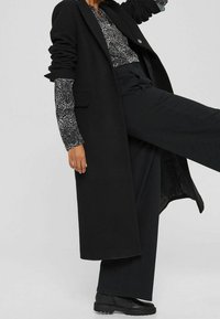 Personne portant un long manteau noir, un pantalon noir, des chaussures noires et une chemise à motifs noir et blanc, levant une jambe vers l'avant.