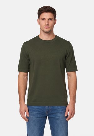 Mężczyzna w ciemnozielonej koszulce z krótkim rękawem i niebieskich dżinsach, stojący na tle jasnego, jednolitego tła.