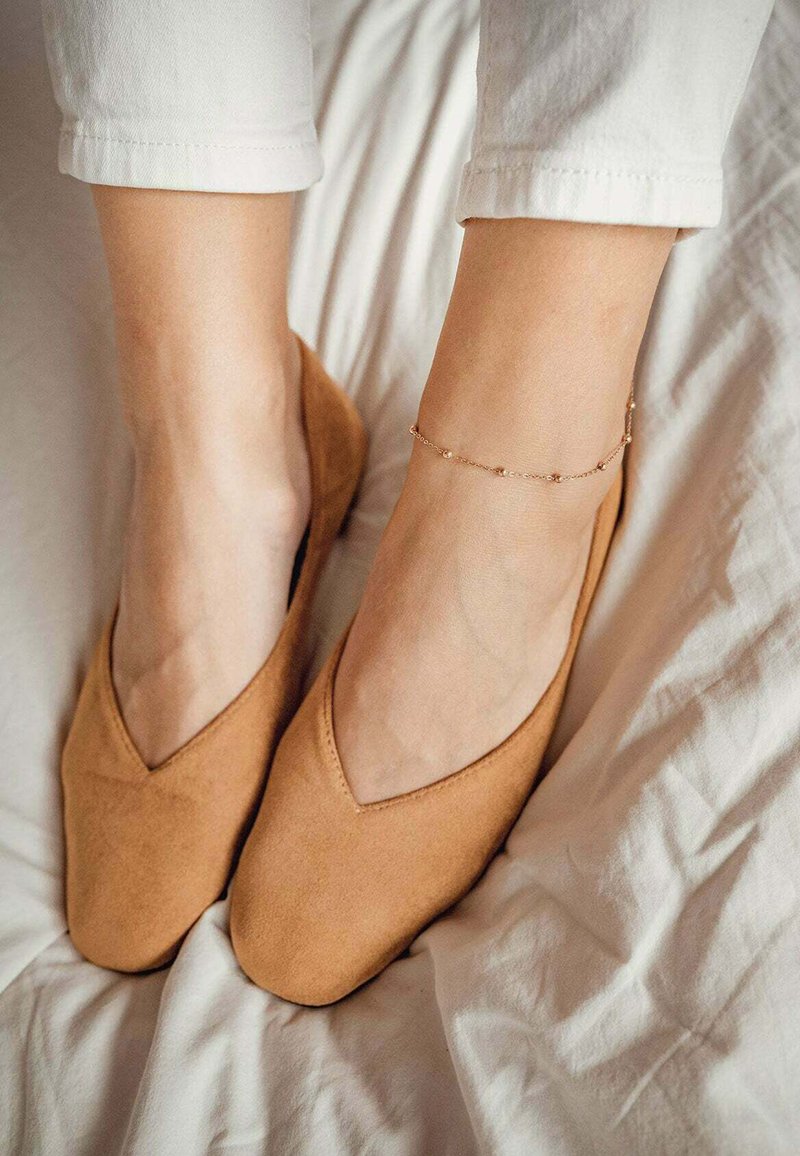 Bailarinas de ante color beige con un diseño en forma de V puntiaguda, situadas sobre una ropa de cama blanca. Un delicado tobillo dorado adorna el tobillo.