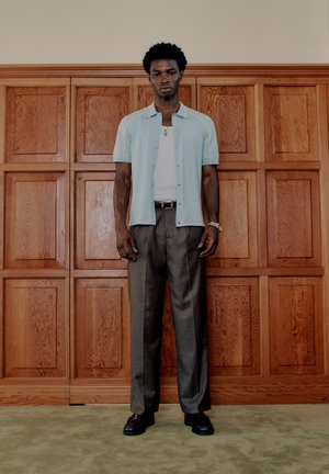 Un homme debout devant un mur en panneaux de bois, portant une chemise à manches courtes bleu clair par-dessus un débardeur blanc, un pantalon gris et des chaussures foncées.