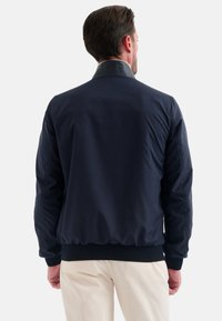 Mörkblå bomberjacka med en slät yta, ribbade ärmslut och svart krage. Designen har en avslappnad passform med elastisk nederkant.