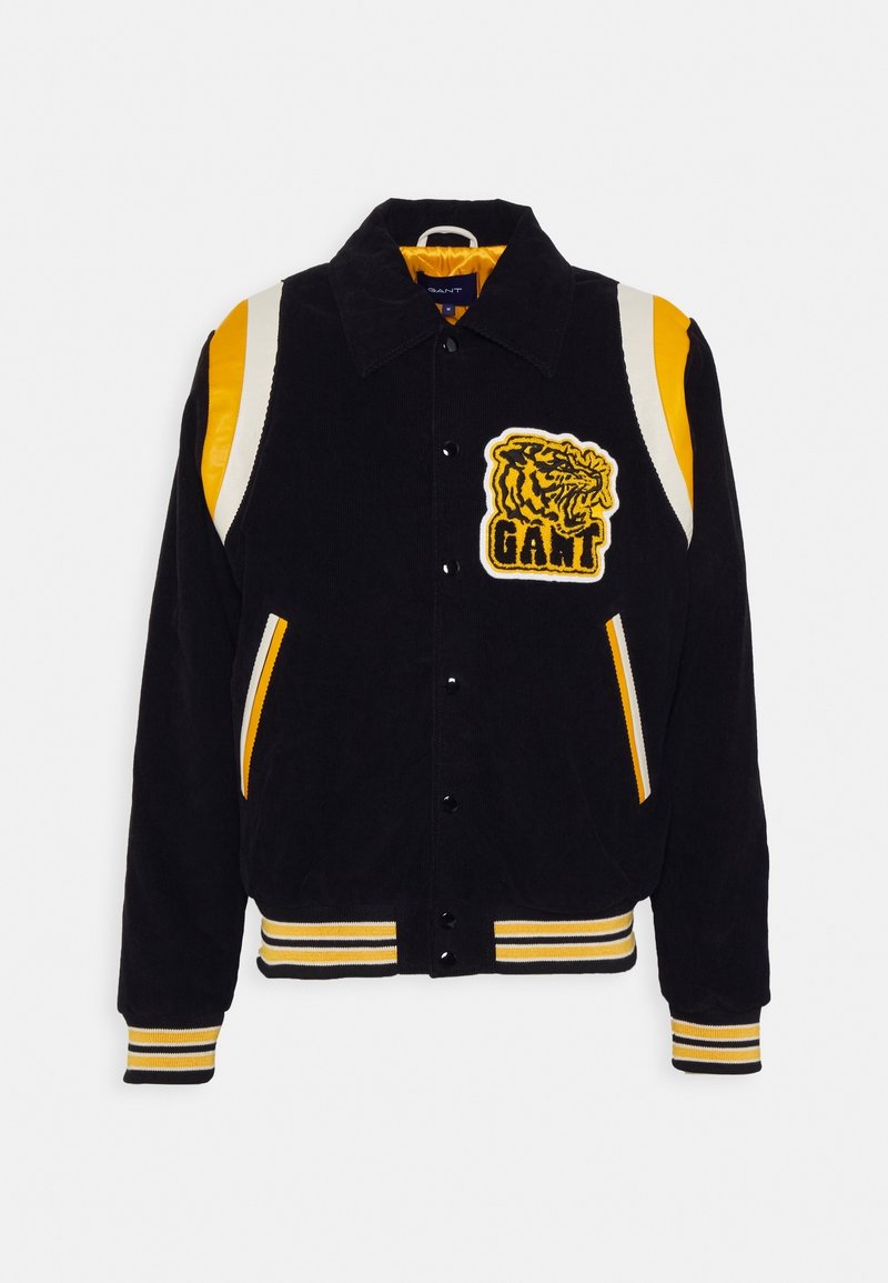 Veste en velours côtelé noir avec des accents rayés jaunes et blancs. Présente un logo de tigre et une fermeture à boutons-pression, avec des poches et des poignets côtelés.