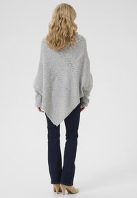 Pelēka adīta pončo ar teksturētu rakstu, kas iezīmē trīsstūrveida formu un brīvi karājošām piedurknēm. Savienots ar tumši ziliem platām džinsām un bēšiem zābakiem.