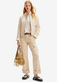 Giacca in suede beige con zip, camicia bianca, pantaloni cargo beige e sandali neri con cinturini blu. Tiene una borsa a tracolla marrone.