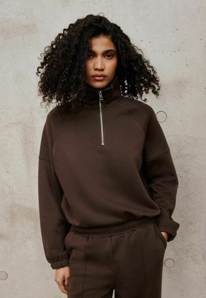 Giovane donna con capelli ricci che indossa una felpa marrone con zip e pantaloni coordinati, in piedi contro un muro di cemento semplice.