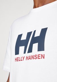 White cotton t-shirt featuring a large navy "HH" logo and "HELLY HANSEN" in red. Round neckline and short sleeves. Smooth texture.
