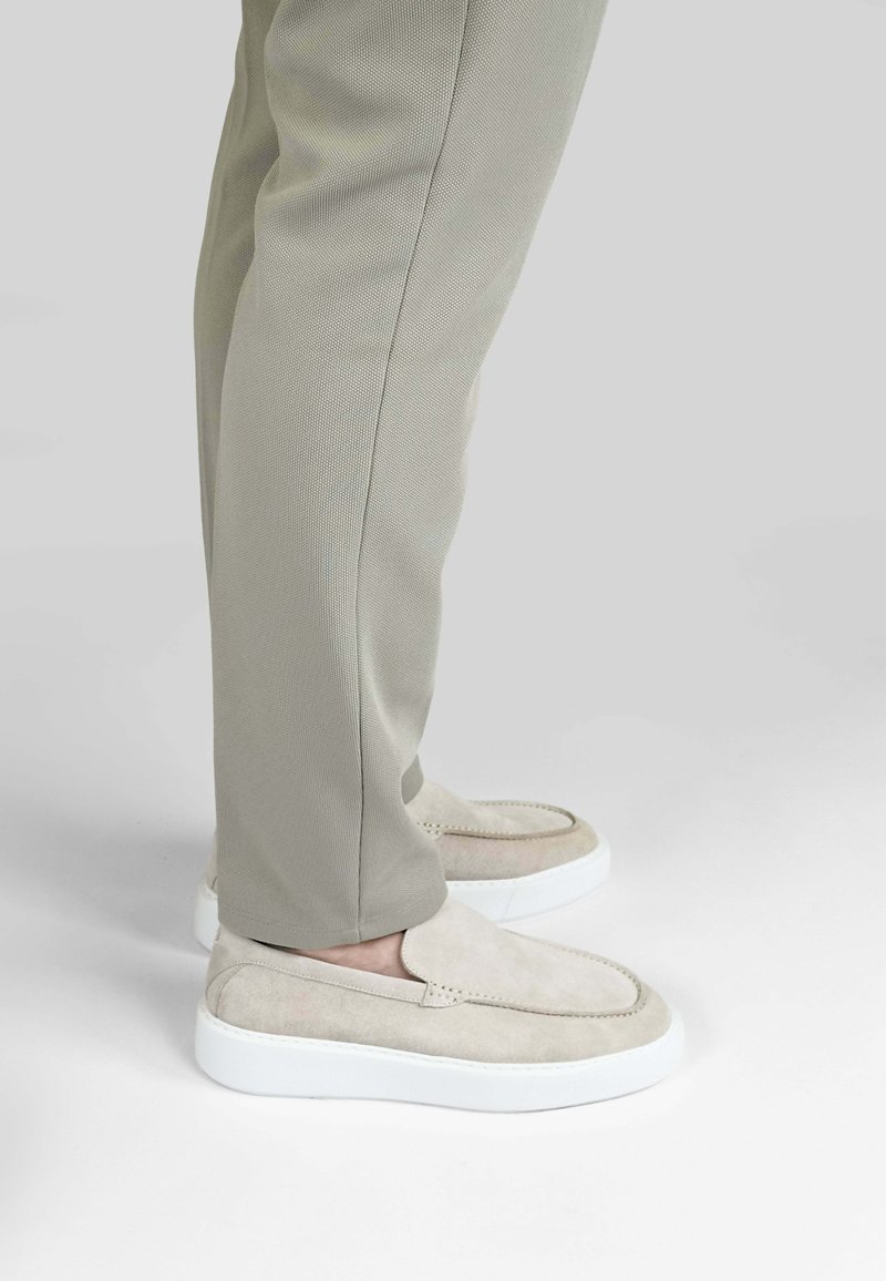 Lichtbeige suède instapschoenen met witte rubberen zolen, met een ronde neus en minimale stikseldetails, gedragen met getailleerde beige broeken.