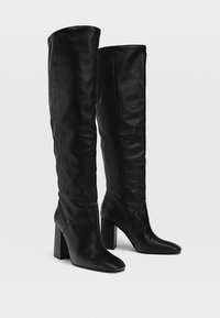 Une paire de bottes noires en cuir haute avec des talons block et des pointes pointues, se tenant côte à côte sur un fond uni.