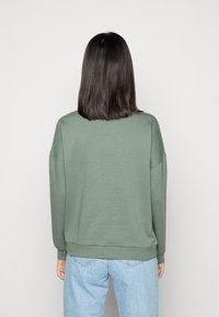 Felpa verde, oversize, con spalle cadenti, polsini e orlo a costine. Indossata sopra jeans azzurri chiari, con texture liscia e design casual.