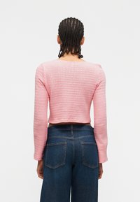 Άτομο με πλεγμένα μαλλιά, φοράει ροζ μακρυμάνικη μπλούζα με υφή και σκουρό μπλε τζιν ψηλόμεσο, γυρισμένο προς τα πίσω πάνω σε απλό φόντο.