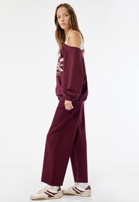 Burgundin ylisuurimallinen collegepaita valkoisella graafisella painatuksella, yhdistettynä leveälahkeisiin burgundin värisiin housuihin ja beigeihin tennareihin, joissa on maroonit yksityiskohdat.