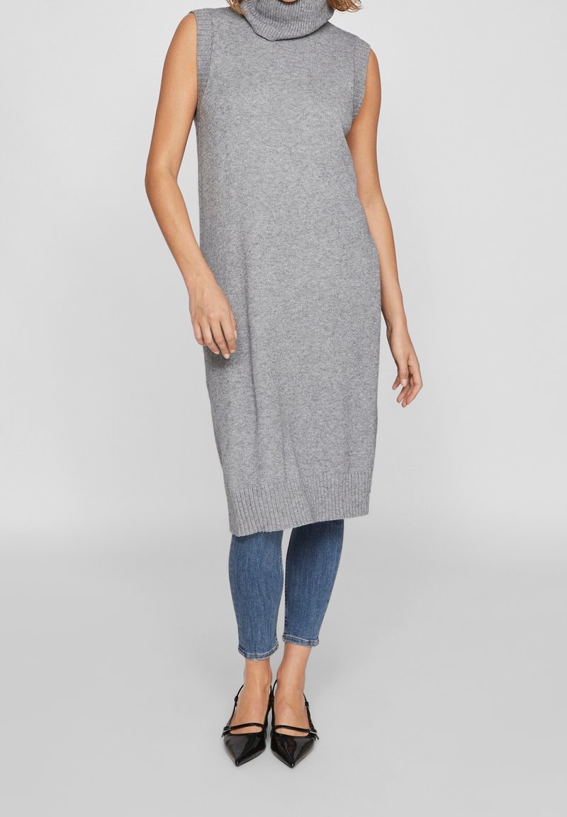 Grå ärmlös tubklänning i mjukt stickat material, knälång med ribbad nederkant, kombinerad med figurnära blå jeans och svarta spetsiga klackskor.