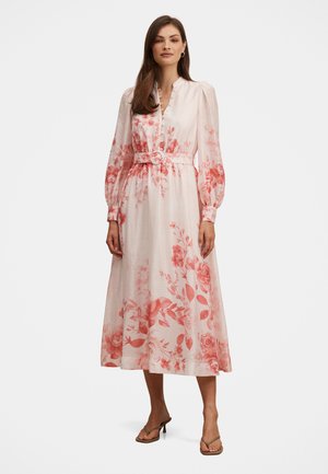 Nő hosszú ujjú, öves, fehér midi ruhában, rózsaszín virágmintákkal, mezítláb áll sima háttér előtt.