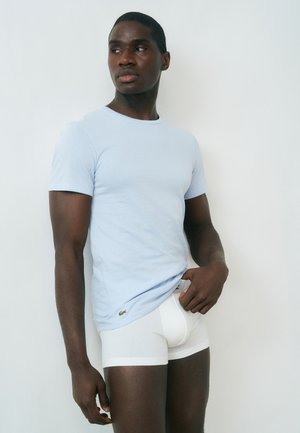 Mężczyzna w jasnoniebieskim t-shircie i białych bokserkach, stojący przy białej ścianie, patrzący na bok, z jedną ręką poprawiającą koszulkę.