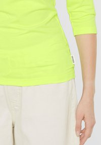 Neon-gelbes Langarmshirt mit einem taillierten Design und einem kleinen weißen Etikett mit der Aufschrift "SENSES" nahe dem Saum, kombiniert mit beigen Hosen.