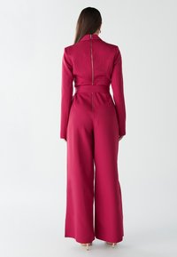 Prilegajo globoko bordo kombinezon z visokim ovratnikom, zapiranjem na zadrgo in širokimi hlačnicami. Tkanina ima gladko teksturo, ki poudari silhueto.