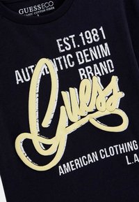 T-shirt in cotone organico nero con testo stampato in bianco e giallo, tra cui "Guess" in un grande carattere corsivo e altri dettagli del marchio.