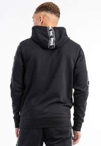 Black cotton hoodie featuring a large hood, ribbed cuffs, and hem. Notable branding tape along the back and sleeves, smooth texture.