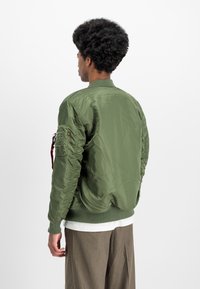 Olive green bomber jacket made of shiny nylon. Features a zippered pocket on the left sleeve and ribbed cuffs and hem.