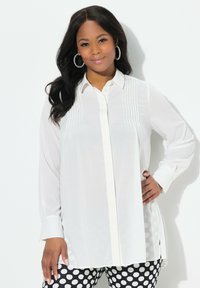 Witte blouse met lange mouwen, met een knoopsluiting aan de voorkant, verticale plooien op de borst en zijsplitten, gecombineerd met zwart-witte polkadotbroek.