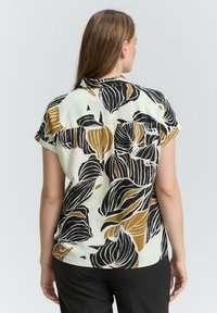 Korte mouwblouse met een crèmekleurige basis en zwarte en mosterdkleurige bladvormen, knoopdetails aan de achterkant en een relaxte pasvorm.