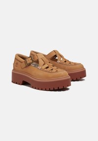 Sapatos de couro castanho com um design trançado, lados abertos e uma sola de borracha vermelha robusta. Apresentam uma tira com uma fivela de metal para ajuste.
