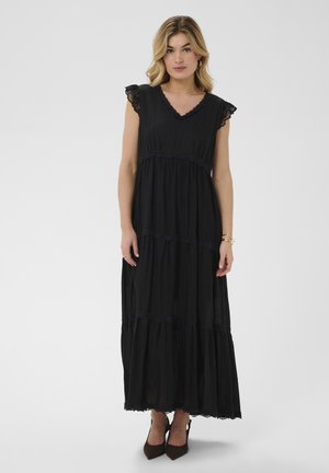 Femme aux cheveux blonds portant une robe maxi noire sans manches avec un bord en dentelle et des talons noirs pointus, debout devant un fond uni.