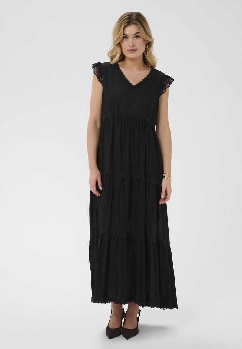Femme aux cheveux blonds portant une robe maxi noire sans manches avec un bord en dentelle et des talons noirs pointus, debout devant un fond uni.