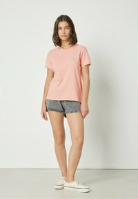 Pink cotton t-shirt with a round neckline and short sleeves, featuring a small green logo on the front. Paired with gray denim shorts.