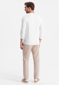 Mann mit dem Rücken zur Kamera, der ein weißes Langarmshirt, beige Hosen, beige Sneaker und ein Perlenarmband am linken Handgelenk trägt.
