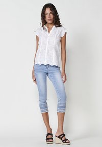 Blouse blanche brodée avec des manches courtes à volants, associée à un jean court bleu clair avec des coutures décoratives et des ourlets effilochés. Sandales à talons compensés noires.