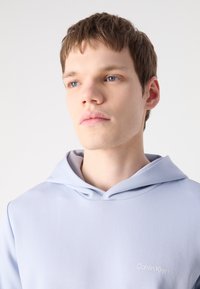 Svijetloplava hoodie izrađena od glatkog materijala s mekanom teksturom. Ima kapuljaču s vezicom i mali bijeli logo na prsima.