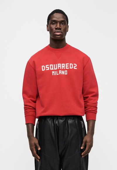 Hombre con sudadera roja DSQUARED2 Milano y pantalones negros, de pie frente a un fondo blanco liso, mirando hacia adelante con los brazos relajados.