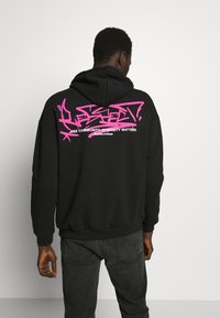 Svart hoodie med frontficka i kängurustil och rosa graffitistil text på baksidan. Har en dragkedja i huvan och ribbade ärmslut.