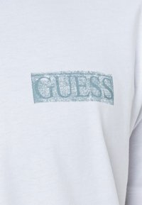 Camiseta de algodón blanca con un estampado desgastado del logo "GUESS" en color azul claro y forma rectangular. Textura suave y ajuste casual.