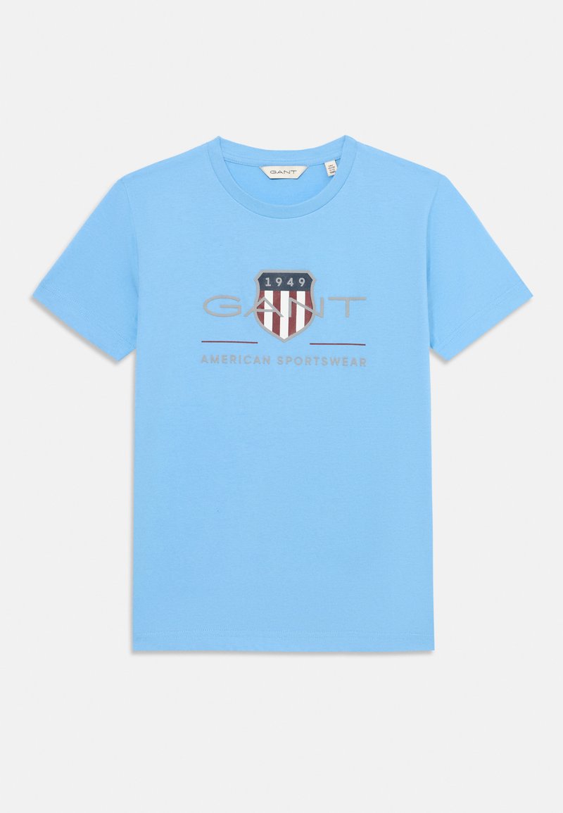 Gaiši zila kokvilnas t-krekls ar centrā izvietotu grafiku, kurā redzams vairoga dizains, teksts "GANT" un "AMERICAN SPORTSWEAR", kā arī gads "1949".