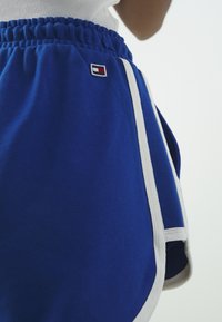 Modré sportovní šortky vyrobené z měkké tkaniny, s bílým lemem, bočními kapsami a malou nášivkou loga na přední straně. Viditelný elastický pas.