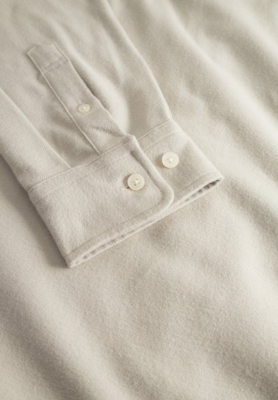 Chemise à manches longues gris clair avec un col rabattu, ornée de deux boutons blanc crème et de tissu doux et texturé.