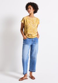 Top corto de mangas cortas con estampado de zigzag amarillo, combinado con jeans de pierna ancha en azul claro y sandalias planas marrones. Estilo casual, ajuste relajado.