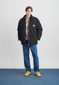 Joven vestido con una chaqueta de plumas negra, sudadera gráfica rosa, jeans azules y zapatillas amarillas, de pie sobre una alfombra azul.