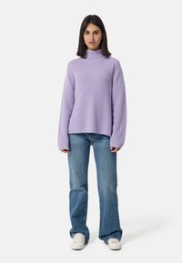 Pull à col roulé rayé de couleur lavande, avec une coupe ample, associé à un jean flare bleu et des baskets blanches, se tenant sur un fond uni.