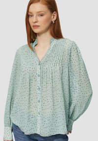 Jasnozielona bluzka w czarne kropki, z plisowanym przodem, bluzkami z marszczeniami i zapięciem na guziki. Okrągły kołnierz i miękka, zwiewna tekstura materiału.