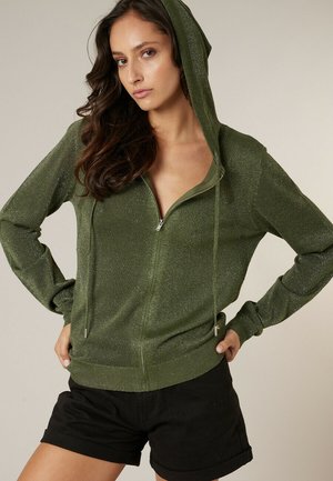 Vrouw draagt een glinsterende olijfgroene hoodie met ritssluiting en capuchon omhoog, met zwarte korte broek, staand met handen in de zij tegen een effen achtergrond.