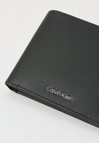Black textured wallet with a smooth finish, featuring a silver "Calvin Klein" logo. Compact design with clean edges.