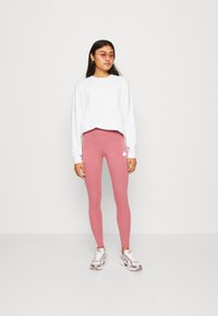 Leggings cor-de-rosa e um suéter longo branco, com sapatos esportivos com acabamentos metálicos, de pé contra um fundo branco simples.